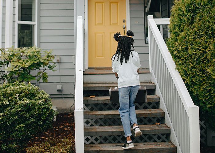 Person walking up stairs to a house with a yellow door, holding a briefcase; evokes mystery theme.