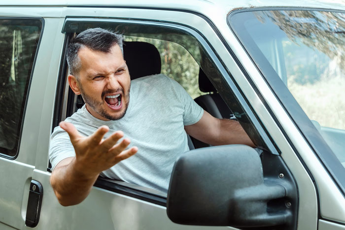 Man in a van yelling with frustration through an open window on a driveway.