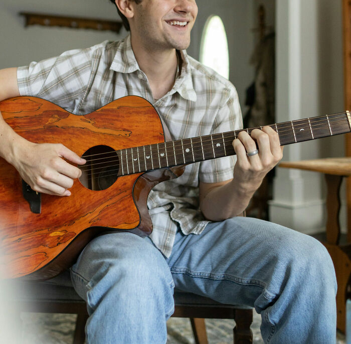 Man playing guitar, wearing a plaid shirt and jeans, smiling indoors; a non-sexual thing men do that attracts women.