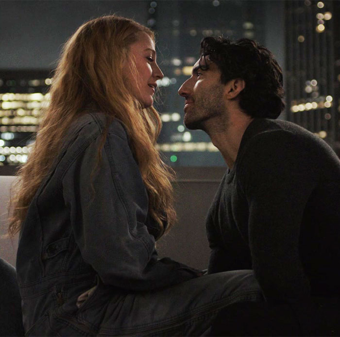 Two people in an intimate moment on a rooftop at night, with city lights in the background.