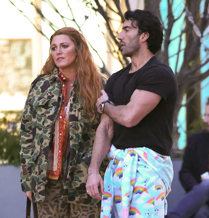 Two people stand outdoors; one in a camo jacket, the other in black with a colorful garment.