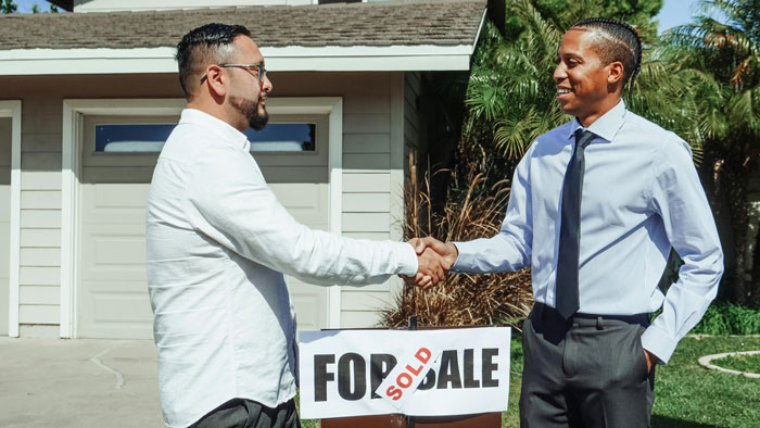 Two men shaking hands in front of a house with a "Sold" sign, discussing a mortgage agreement.