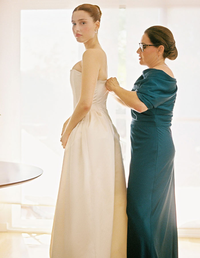 Bride in a dramatic wedding dress, assisted by a woman, showcasing elegance and style.