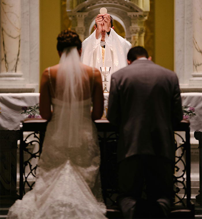 A bride and groom kneeling at the altar during a wedding ceremony, with a priest holding a host.