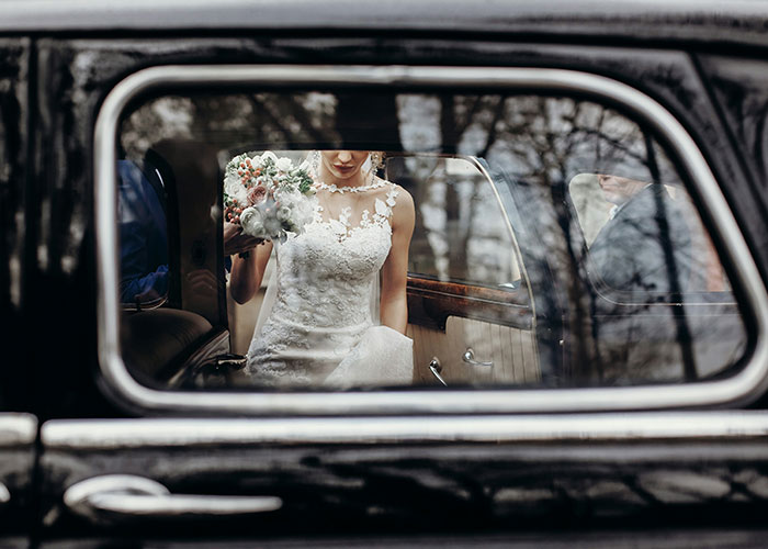 Bride in a lace wedding dress holding a bouquet, seen through a car window, symbolizing newlyweds' transportation plans.