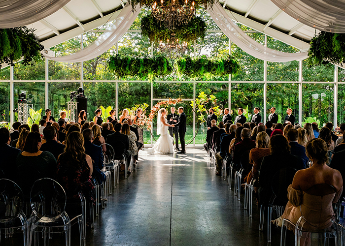 Newlyweds at an indoor wedding ceremony with guests seated, featuring elegant decor and lush greenery.