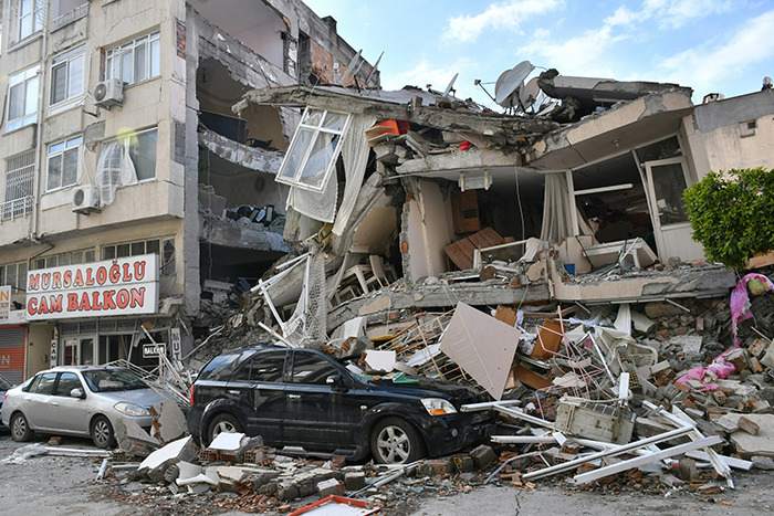 Earthquake aftermath showing collapsed building and damaged cars, highlighting challenges in predicting such disasters.