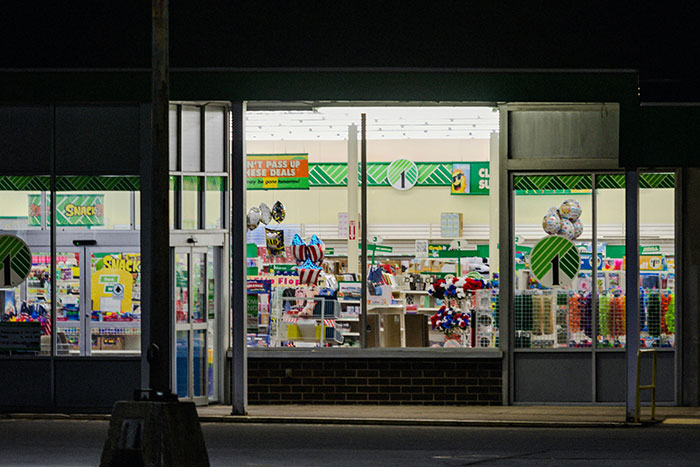 Retail store exterior at night, brightly lit interior displays snacks, balloons, and supplies.