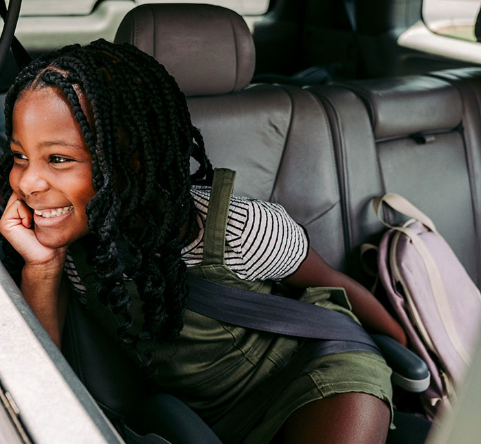 A girl smiling in the backseat of a car, wearing a striped shirt and green overalls, with a backpack beside her.