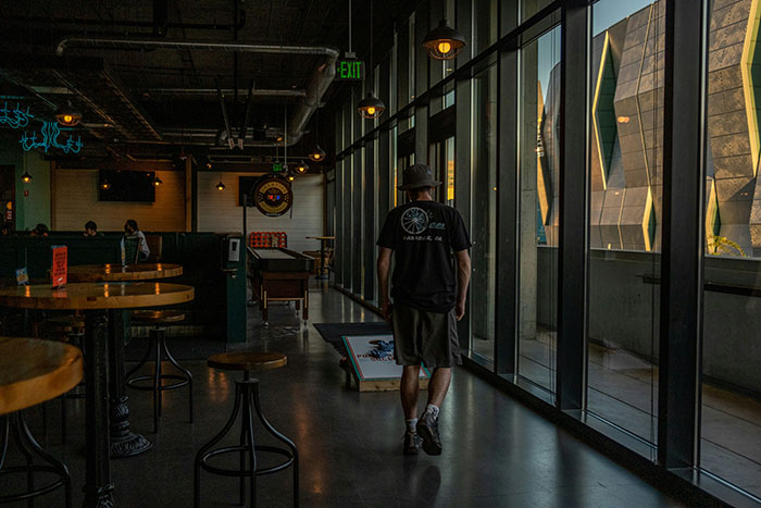 Retail worker walks through a dimly lit cafe with stools and a game board near large windows.