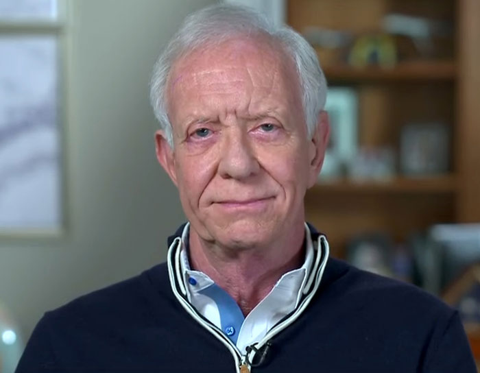 Man in a blue shirt and dark sweater discusses a plane crash theory involving fewer visible ground lights.