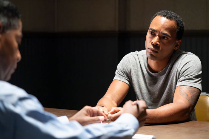 Actor Maurice Marvel in a gray shirt sitting at a table, likely from the cast of "High Potential," looking focused in a dramatic scene.