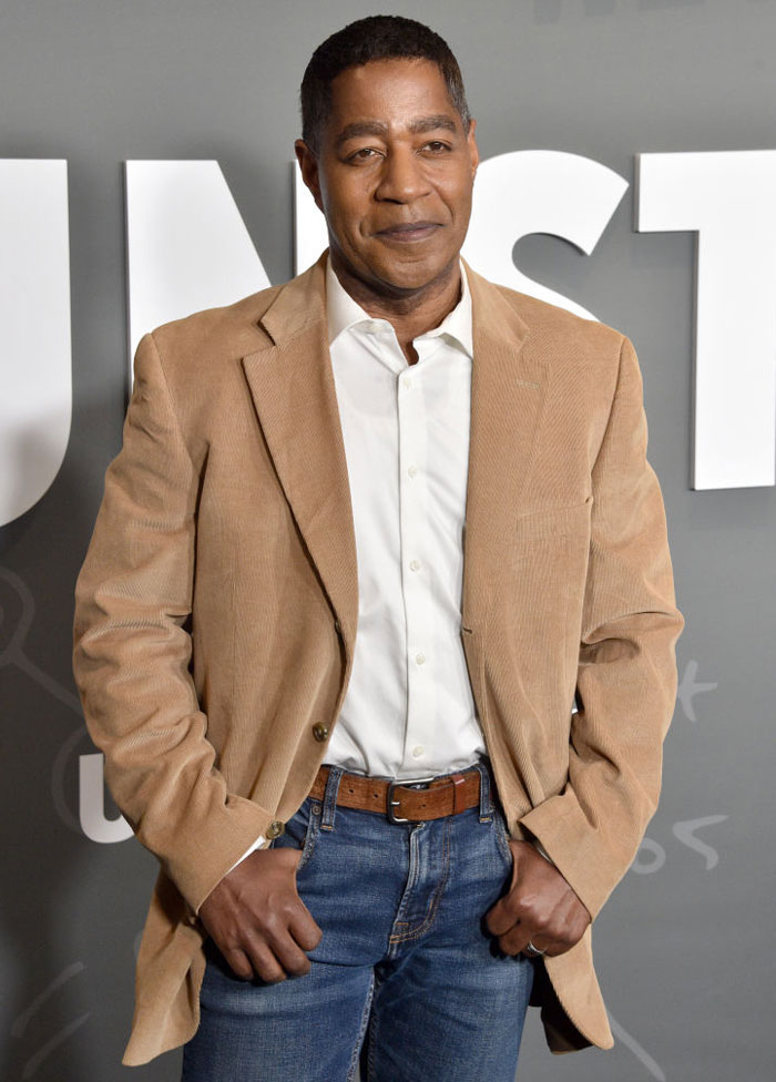 Actor Merrick McCartha in a tan blazer and jeans, posing in front of a grey background, part of the "High Potential" cast.