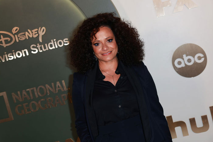 Judy Reyes with curly hair poses in front of a Disney Television Studios backdrop, representing the cast of High Potential.