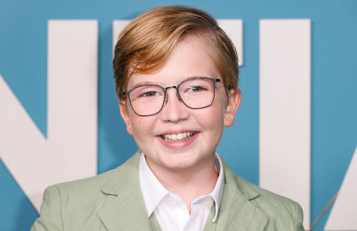 Matthew Lamb, young cast member of High Potential, wearing glasses and a light green suit, smiling against a blue background.