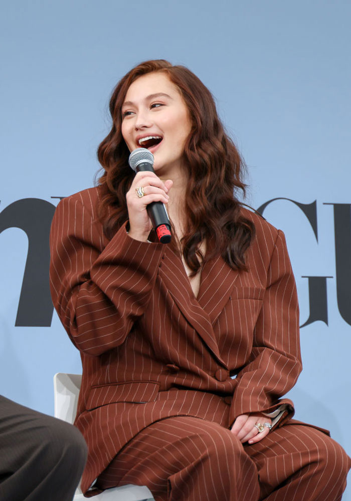 Lola Tung from "The Summer I Turned Pretty" in a pinstripe suit, speaking into a microphone at an event.