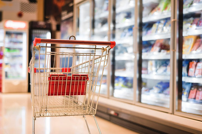 An empty shopping cart in a grocery store aisle with freezer section on the right.