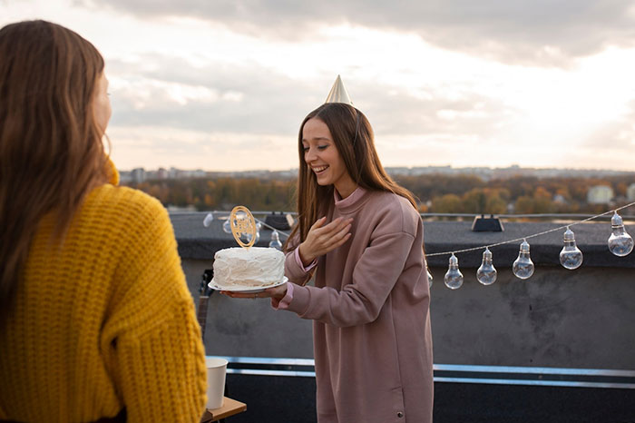 Woman holding a cake while smiling at a rooftop gathering, wearing a party hat, with a background of lights and trees.