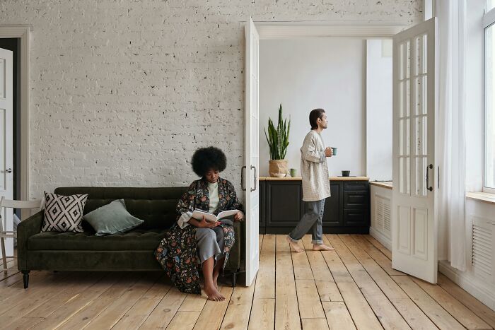 Living cheap tip: Cozy living room with a person reading on a sofa and another walking with a mug in a bright kitchen.