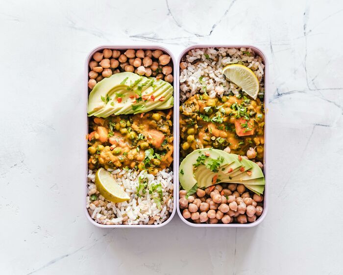 Budget-friendly meal of rice, chickpeas, lentils, avocado, and lime in two containers on a white surface.