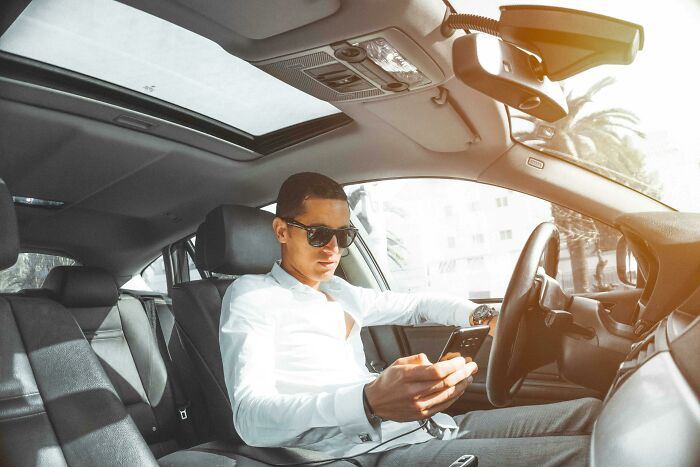 Man in white shirt and sunglasses sitting comfortably in a car, browsing his phone.