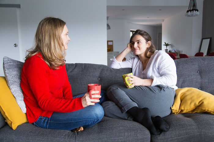 Two women sharing tips for living cheap while comfortably seated on a cozy sofa, each holding a mug.