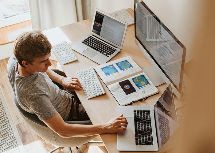 Person at a desk with multiple laptops and a notebook, working on cheat codes for life in a productive setup.