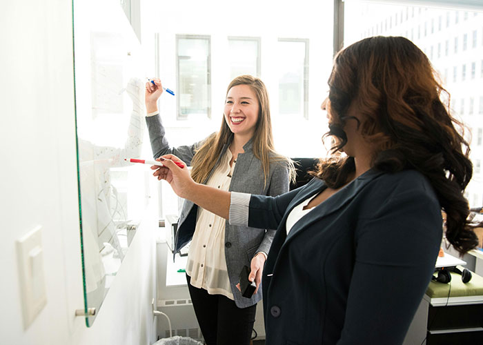 Two women discussing life cheat codes at a whiteboard, smiling in a bright office setting.