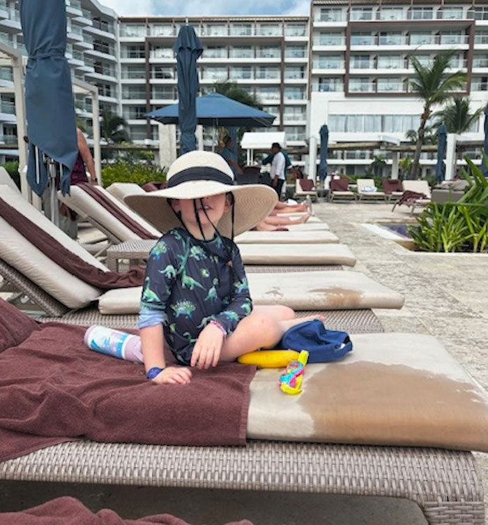 Child in a sun hat on resort lounge chair by poolside in Cancun, surrounded by vacationers.