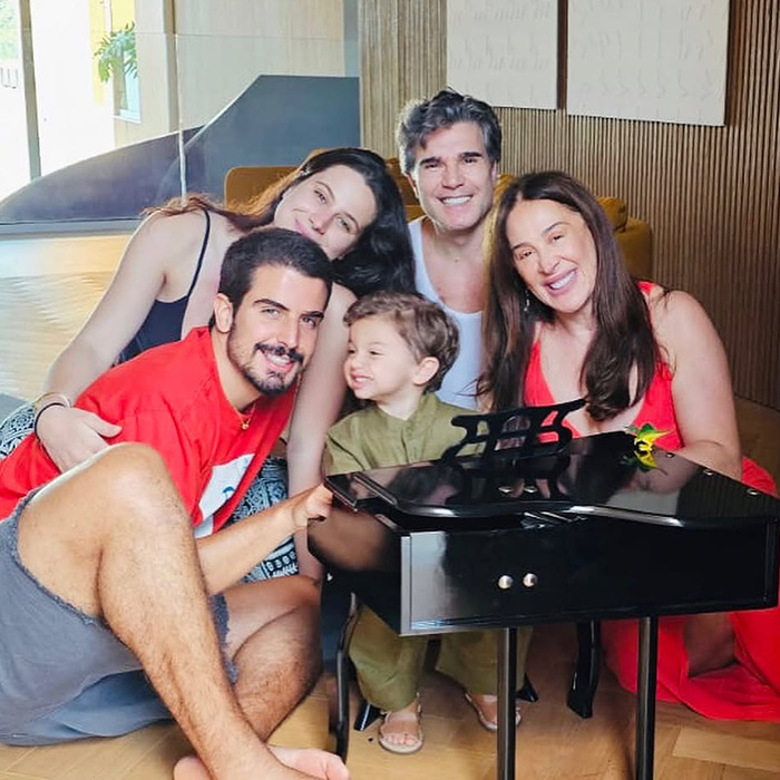 Claudia Raia and family gathering around a small black piano, with members smiling and interacting warmly indoors.