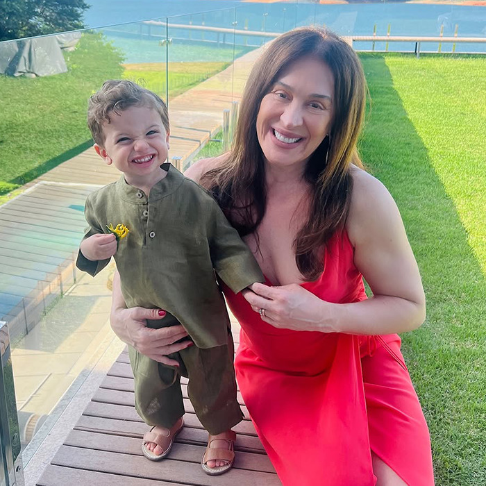 Claudia Raia and toddler smiling outdoors on a sunny day, green lawn and pathway in background.