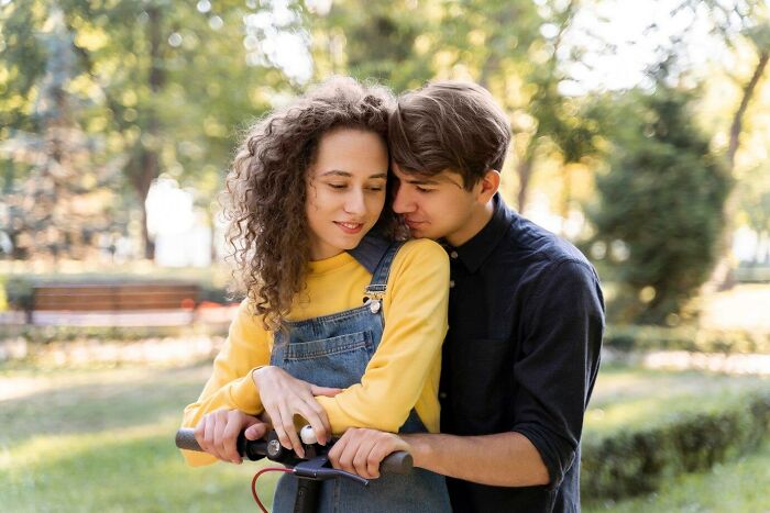 A young couple shares a tender moment on a scooter in a park, surrounded by greenery.