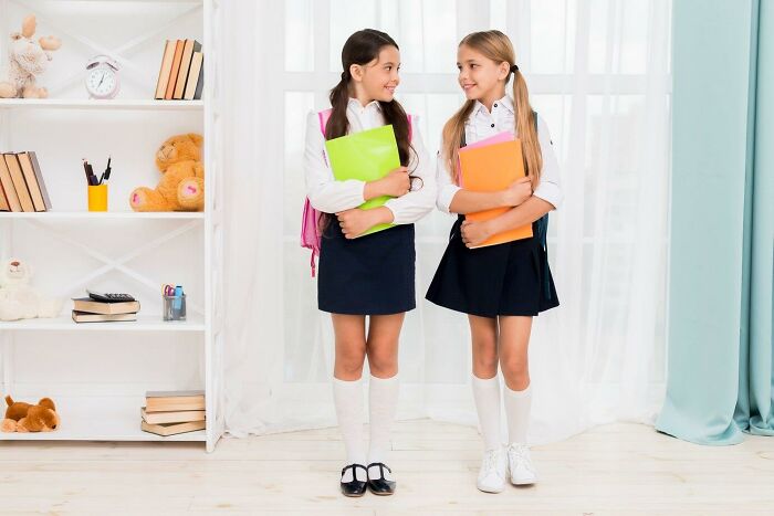 Two schoolgirls in uniform, holding folders, standing and smiling at each other in a room with a bookshelf.