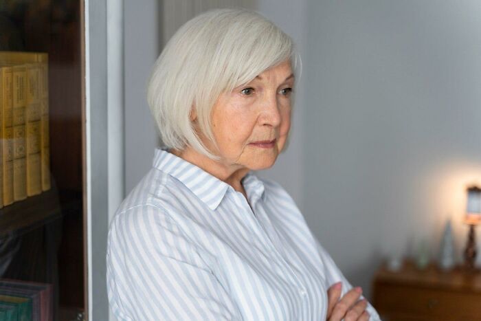 An older woman in a striped shirt looking thoughtful, standing indoors near a bookshelf.