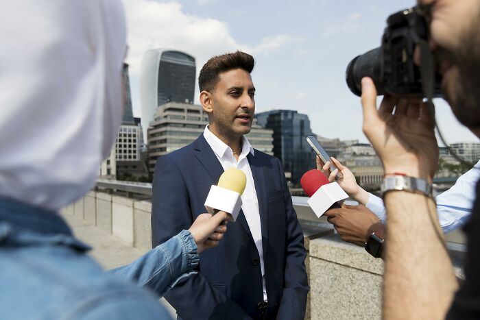 Man in a suit speaking to reporters, multiple microphones around him, skyscrapers in the background, discussing a major human history scam.