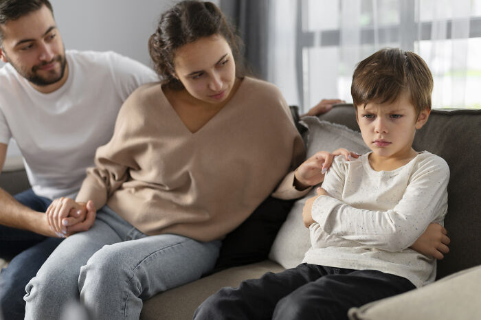 A concerned couple sits with a sad child on a couch, highlighting parenting issues.