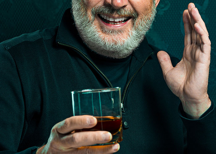 Man with a beard holding a glass of whiskey, smiling and gesturing, symbolizing sharing dark family secrets.