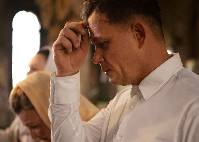 A man in deep thought indoors, dressed in a white shirt, reflecting on family secrets.