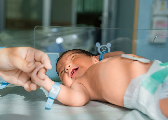 Newborn baby holding an adult's finger, wearing a hospital bracelet, under soft lighting.