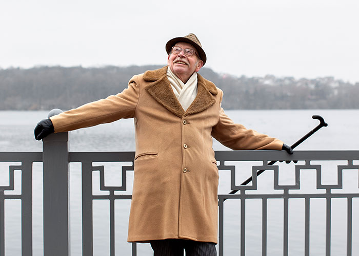 Elderly man in a coat and hat, smiling by a lake, holding a cane.