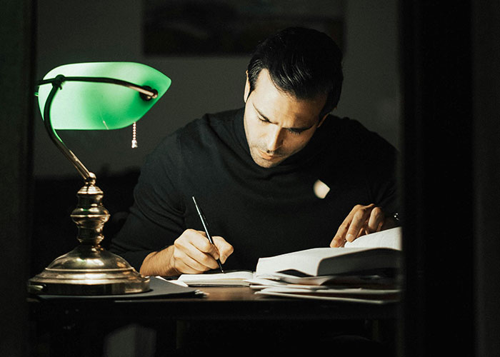 Man writing under a green desk lamp, possibly reflecting on darkest family secrets in solitude.