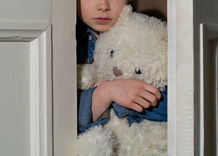 A child leans out from behind a door, holding a teddy bear, representing secretive family themes.