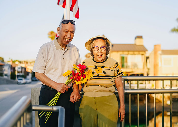 Elderly couple holding flowers, smiling on a sunny day, representing family secrets theme.