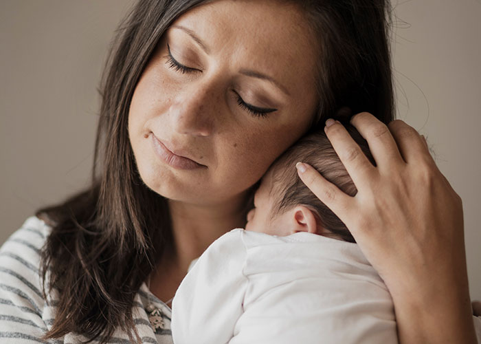 A woman holds a baby, eyes closed, conveying warmth and protection; themes of family secrets explored online.