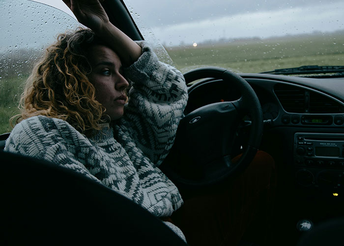 Woman in a car, looking unsettled with her hand on her forehead, during a rainy day.