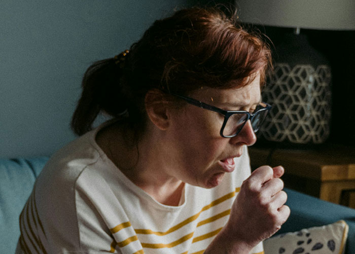 Woman with unsettling expression, coughing into her hand while seated, wearing glasses and a striped shirt.