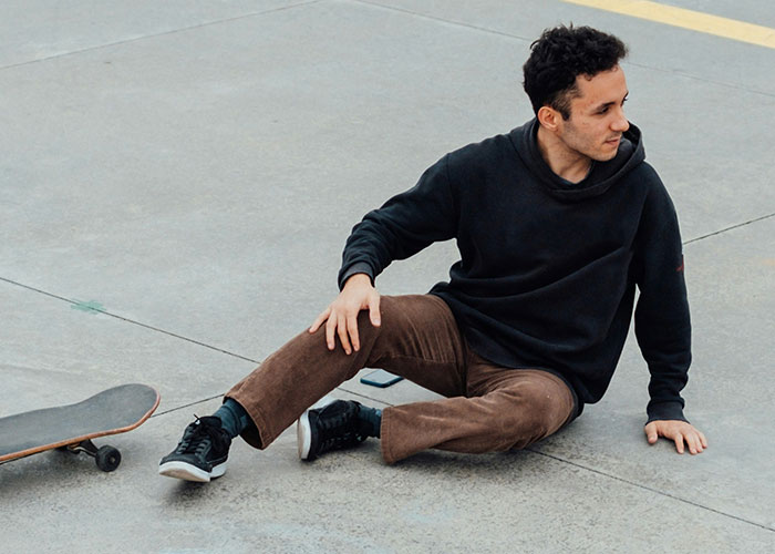 Man sitting on the pavement beside a skateboard, wearing a black hoodie and brown pants.