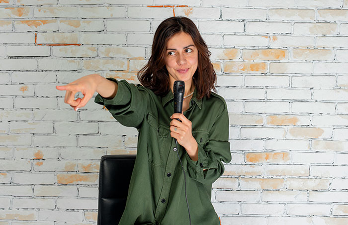 Woman smiling with a microphone, casually dressed, pointing playfully against a brick wall background.