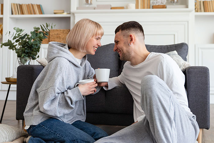 Couple sitting on the floor, smiling and drinking coffee together in a cozy living room, representing adorable partner moments.