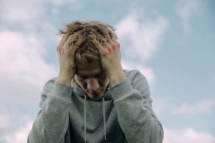 Person in a gray hoodie outside, expressing stress or contemplation, with hands on their head against a cloudy sky.